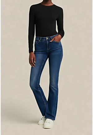 Henkilö, jolla on musta pitkähihainen paita, siniset korkeavyötäröiset farkut ja valkoiset kengät, seisoo käsi taskussa.