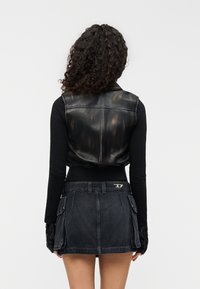 Μαύρο δερμάτινο κοντό γιλέκο με γυαλιστερή υφή, σε συνδυασμό με εφαρμοστή μαύρη μακρυμάνικη μπλούζα και σκουρόχρωμη μίνι φούστα από τζιν με τσέπες cargo.