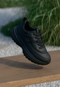 Svarta sportskor med en texturerad ovansida, tjock gummisula och framträdande mönster. Har snörning och en dragflik för enkel påtagning.