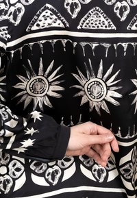 Schwarzer Stoff mit auffälligen weißen Mustern von Sonnenmotiven, geometrischen Formen und floralen Designs; Langarm mit gerafftem Manschetten-Detail.
