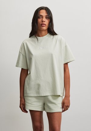 Femme aux longs cheveux foncés portant un t-shirt beige clair oversize et un short côtelé, debout devant un fond gris uni.