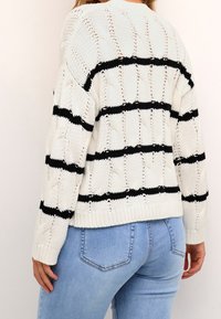 Pull en maille blanche avec des rayures horizontales noires, présentant une texture en maille torsadée et un ourlet côtelé, associé à un jean bleu clair.