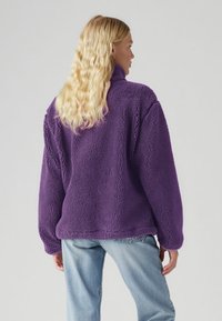 Lila polár pulóver magas gallérral, texturált, bolyhos felülettel és kényelmes szabással. Világoskék farmerral viselve.