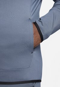 Blue athletic top with long sleeves, featuring a fitted design, side slits, and black accents at the cuffs and hem. Smooth texture.