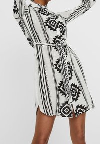 Vit klänning med svarta geometriska mönster, långa ärmar och ett knytbälte i midjan. Har sidoflikar och en kragdesign.