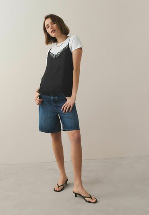 Femme portant un t-shirt blanc sous un caraco noir à dentelle, un short Bermuda en jean et des sandales à talons noires sur un fond clair.