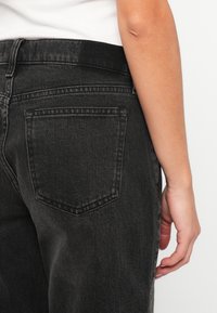 Jean en denim noir à taille haute, avec une poche arrière visible et une texture légèrement décolorée, associé à un haut blanc simple et ajusté.