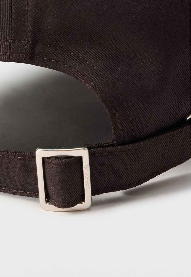 Cappello in tessuto marrone con fibbia metallica regolabile. Il materiale ha un aspetto strutturato con dettagli di cuciture visibili.