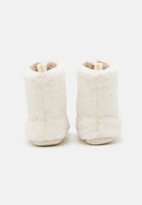 White plush ankle boots with soft, furry texture, beige lining, and a flat sole. Includes a fabric loop tag for easy wearing.