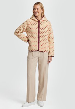 Smilende kvinde iført beige hættejakke med hjertermønster, beige bukser med brede ben og hvide sneakers, stående mod en ensfarvet baggrund.