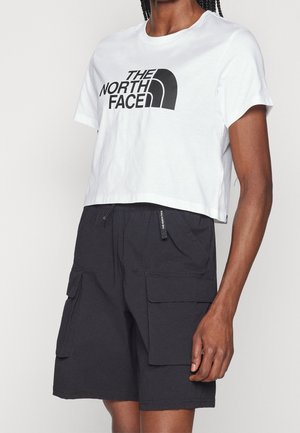 Osoba ubrana w białą krótka koszulkę z logo "The North Face" oraz czarne szorty typu cargo z przednimi kieszeniami z klapami.