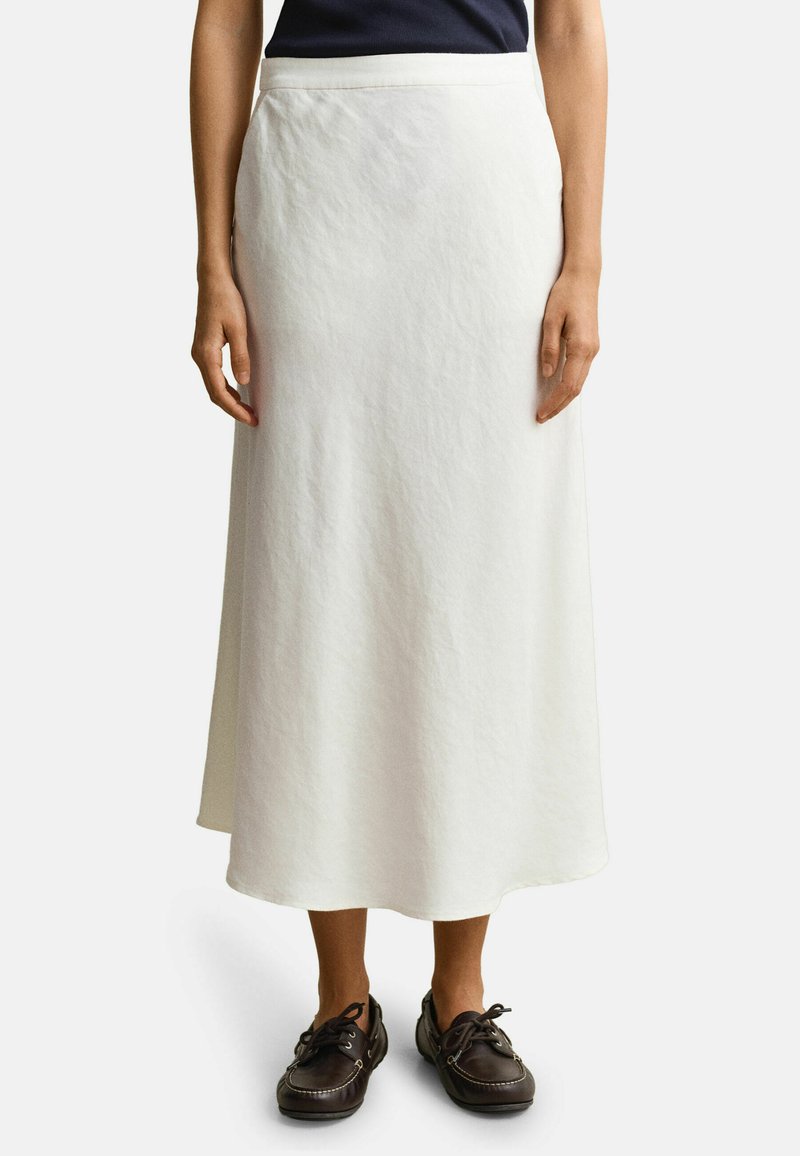 Frau trägt einen weißen Midirock, navy blaues Oberteil und dunkelbraune Schnür-Loafers, steht vor einem einfarbigen Hintergrund.