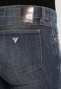 Džíny z denimu s tmavým ombré, které mají koženou nášivku GUESS, kovový trojúhelníkový logo detail a dekorativní cvočky na boku.