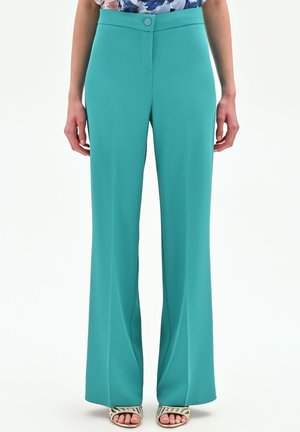 Pantaloni a vita alta verde petrolio a gamba larga con bottone frontale e vita liscia, indossati con sandali dorati aperti con cinturini.