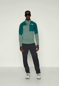 Veste zippée pour homme en teal et gris avec des panneaux texturés. Associée à un pantalon noir et des chaussures de randonnée claires. Lunettes de soleil portées.