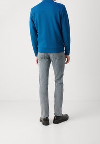 Blue sweatshirt with a high collar, elastic cuffs and hem, paired with light gray jeans and black shoes. Rear view showcases the design and fit.