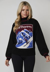 Donna con lunghi capelli biondi che indossa un maglione nero con una scena sciistica di montagne e picchi blu, con la scritta "Val Thorens".