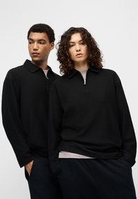 Μαύρο πουλόβερ με φερμουάρ στον γιακά, με υφή πλεξίματος. Και τα δύο μοντέλα φορούν ταιριαστά εξωτερικά ρούχα με άνετη εφαρμογή και μακριά μανίκια.