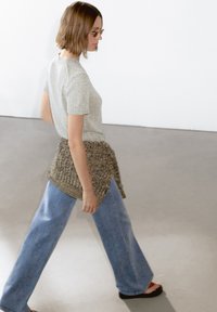 Maglione grigio chiaro a maniche corte, indossato sopra un ampio wrap in maglia marrone testurizzato, abbinato a jeans in denim blu a gamba larga e sandali neri.