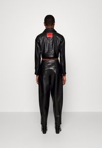 Zwarte neplederen cropped jas met een rood "HUGO" label op de achterkant, gecombineerd met loszittende zwarte neplederen broeken.