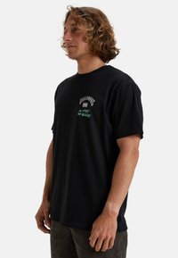 Black cotton t-shirt with short sleeves, featuring front embroidery in orange and green text. Relaxed fit, rounded neckline, smooth texture.