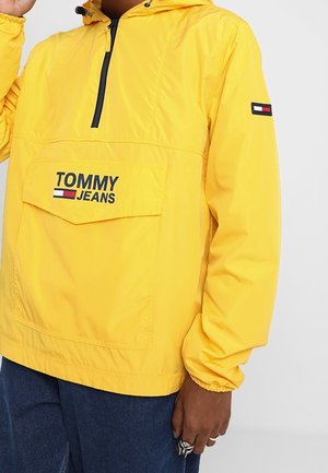 Żółta kurtka z kapturem Tommy Jeans z przednią kieszenią i czarnym suwakiem, noszona z ciemnoniebieskimi spodniami, z widocznym lewym ramieniem i ręką.