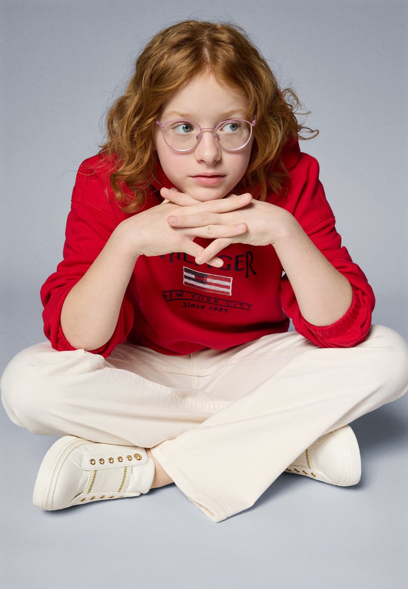 Sweatshirt rouge avec un design graphique, pantalon large crème, baskets blanches avec des accents dorés, et lunettes roses rondes.