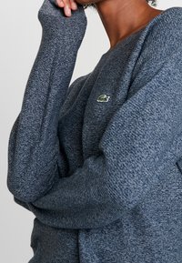 Pull bleu foncé avec un tricot texturé, coupe ajustée et manches longues. Présente un petit logo vert sur la poitrine.