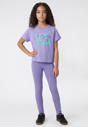 Meisje met krullend haar, gekleed in een paarse "Live & Dream" T-shirt en legging, met zwarte sneakers, staand met een hand op haar heup.