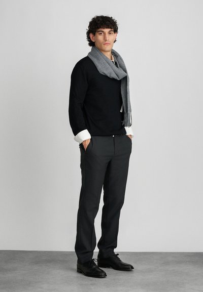 Chemise noire à manches longues, poignets blancs, écharpe grise, pantalon gris et chaussures noires. Le mannequin se tient droit devant un fond neutre.