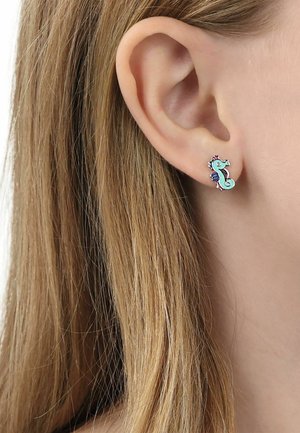 Primo piano di un orecchio che indossa un piccolo orecchino a bottone a forma di cavalluccio marino turchese e rosa, con capelli biondi sullo sfondo.