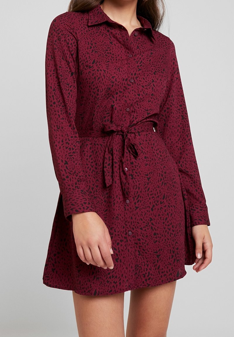 Femme portant une robe bordeaux à manches longues, boutonnée, avec un imprimé léopard noir et une ceinture à la taille, debout devant un fond uni.