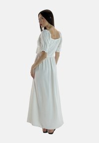 Hvid lang kjole med en tekstureret, smocked bodice og puffærmer. Har et firkantet rygdesign og falder til anklerne. Bæres med sorte hæle.