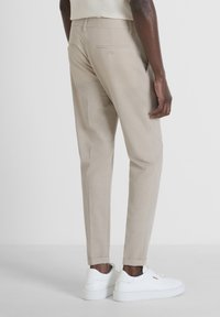 Pantaloni beige su misura con vestibilità slim, dotati di una tasca posteriore con chiusura a bottone e orli cuciti, abbinati a sneakers bianche.