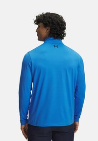 Leichtes Langarmshirt aus leuchtend blauem Stoff mit hohem Kragen, auf dessen Rückseite sich ein schwarzes Logo befindet. Glatte Textur und taillierte Passform.