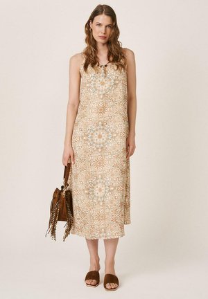 Mujer con vestido midi beige sin mangas y estampado, sosteniendo un bolso marrón con flecos, y llevando sandalias marrones de tacón ancho y abierto, de pie.