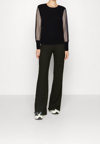 Top nero con maniche in rete trasparente, abbinato a pantaloni a gamba larga verde scuro con texture. Outfit completato da sneaker bianche chunky.