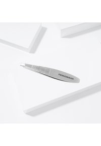 Pinzette Tweezerman inclinate in acciaio inossidabile appoggiate su superfici geometriche bianche, che mostrano la presa texturizzata e il nome del marchio.