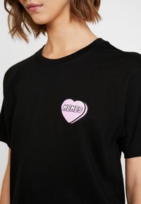 Czarna bawełniana koszulka z różowym graficznym sercem i napisem "MEMES" w białym kolorze na lewej części klatki piersiowej. Krótkie rękawy.