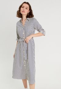 Robe à manches courtes, boutonnée, avec des rayures verticales bleu marine et blanches, deux poches poitrine, taille cintrée avec une ceinture, longueur au genou. Tissu en coton.