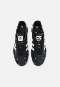 Pár černých tenisek Adidas s bílými pruhy a modrými květinovými vzory na špičkách, viděno zeshora na bílém pozadí.