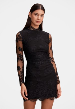 Femme aux longs cheveux foncés portant une mini robe en dentelle noire avec des manches florales transparentes et un col haut, posant devant un fond uni.