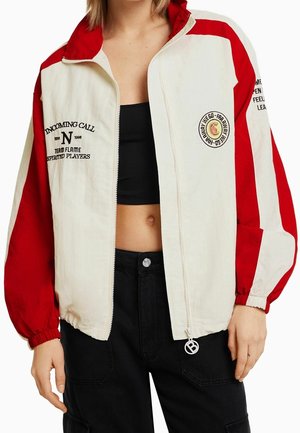 Femme portant une veste zippée blanche et rouge avec un crop top noir et un pantalon noir. La veste présente du texte et un logo circulaire à l'avant et sur la manche.