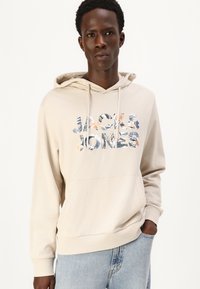 Haut à capuche beige avec une poche kangourou, cordon de serrage à la capuche et un logo "JACK & JONES" coloré arborant des motifs tropicaux. Tissu en mélange de coton doux.