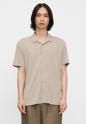 Junge Person mit mittellangen schwarzen Haaren, die ein beiges kurzärmeliges Hemd und braune Hose vor weißem Hintergrund trägt.