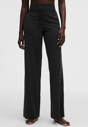 Persona che indossa pantaloni neri a vita alta e gamba larga con coulisse, in piedi a piedi nudi su sfondo chiaro uniforme.