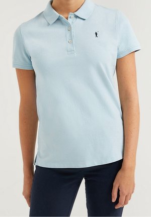 Kobieta w jasnoniebieskiej koszulce polo z krótkim rękawem, z małym haftowanym logo golfisty na lewej piersi, w połączeniu z ciemnymi spodniami.