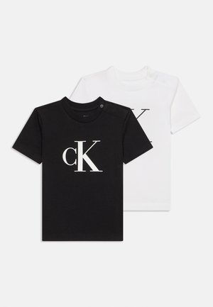 Due magliette a maniche corte per bambini piccoli, una nera e una bianca, ciascuna con un grande logo CK sul davanti e bottoni automatici sulla spalla all'altezza del collo.