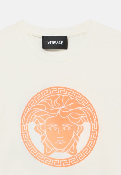 Λευκό βαμβακερό t-shirt με στρογγυλό πορτοκαλί γραφικό ενός προσώπου με διακοσμητικά μαλλιά, περιβαλλόμενο από επαναλαμβανόμενο μοτίβο ελληνι κού κλειδιού. Ορατή ετικέτα Versace.