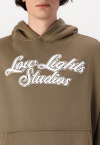 Maslinasto zeleni dukserica s kapuljačom s bijelim izvezenim logotipom "Low Lights Studios". Ima prednji džep i standardni dizajn kapuljače. Glatka tekstura.